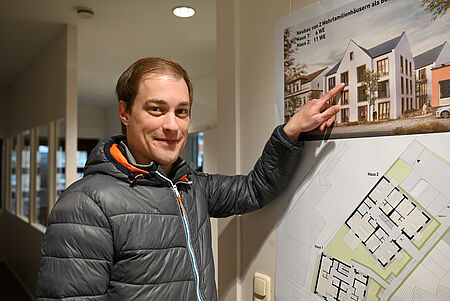 Richtfest_Breite Straße 2025 Ein Mann in grauer Steppjacke steht in einem Innenraum und lächelt in die Kamera. Mit einer Hand zeigt er auf ein an der Wand hängendes Poster, das die Visualisierung und den Grundriss zweier geplanter Mehrfamilienhäuser zeigt. Das Poster enthält eine architektonische Außenansicht sowie die Grundrisszeichnung der Gebäude.