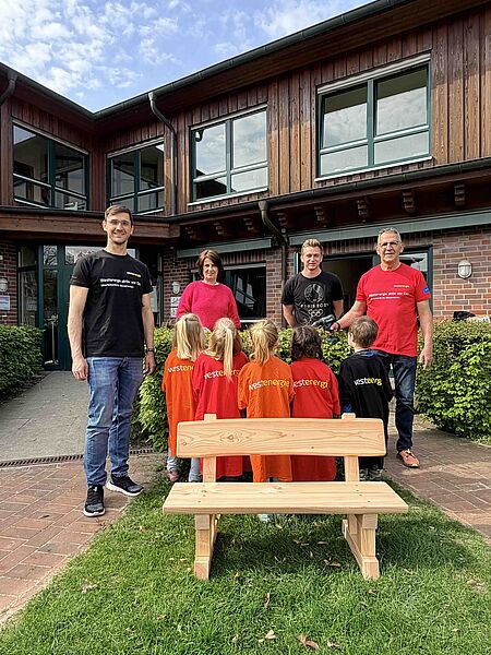 Spende Kita Regenbogen Ein Gruppe von Erwachsenen und Kindern steht vor einem Gebäude mit Holzfassade und großen Fenstern. Die Erwachsenen tragen T-Shirts mit dem Aufdruck „Westenergie aktiv vor Ort“ und halten gemeinsam mit den Kindern, die orange und rote T-Shirts mit dem Aufdruck „Westenergie“ tragen, eine kleine Feier ab. Im Vordergrund steht eine selbstgezimmerte Holzbank, die vermutlich im Rahmen einer Gemeinschaftsaktion entstanden ist. Das Bild vermittelt den Eindruck einer gelungenen Kooperation und eines Gemeinschaftsprojekts.