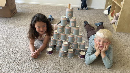 06.08.2025_Abenteuerland Zwei Kinder, ein Mädchen und ein Junge, liegen auf dem Bauch auf einem beigen Teppichboden. Zwischen ihnen befindet sich ein Turm, der aus vielen grauen Pappbechern gebaut wurde. Das Mädchen auf der linken Seite schaut in die Kamera und lächelt. Der Junge auf der rechten Seite stützt seinen Kopf in seine Hände und schaut ebenfalls in die Kamera. Im Hintergrund sind Regale mit verschiedenen Gegenständen und eine Pappschachtel zu sehen.