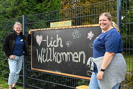 Kita-Fest_26.09.2025 Zwei Mitarbeiterinnen begrüßen Kinder und Eltern neben einer Tafel mit der Aufschrift „Herzlich Willkommen – Schukis 2025“.