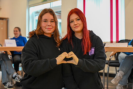 Schulsanitäter_Fortbildung Zwei junge Frauen sitzen nebeneinander, tragen schwarze Hoodies und formen mit behandschuhten Händen gemeinsam ein Herz. Sie blicken in die Kamera, im Hintergrund sind weitere Teilnehmende eines Workshops zu sehen.