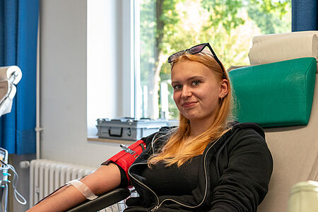 Blutspende_Lengerich Eine junge Frau sitzt entspannt auf einem Spenderstuhl und lächelt leicht in die Kamera. Sie trägt eine schwarze Jacke und hat eine Blutspende-Nadel im linken Arm, der mit einem roten Halter fixiert ist. Im Hintergrund sind ein Fenster mit grünem Ausblick sowie medizinische Geräte zu sehen.