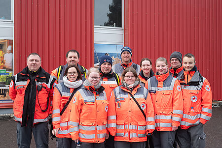 DRK_Karneval Elf Einsatzkräfte des Deutschen Roten Kreuzes stehen in zwei Reihen vor einer roten Gebäudefassade und blicken in die Kamera. Sie tragen orangefarbene Einsatzjacken mit reflektierenden Streifen und DRK-Emblem, teilweise mit Funkgeräten und Mützen. Die Gruppe wirkt geschlossen und einsatzbereit.