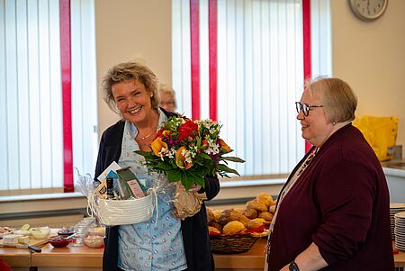 40 Jahre DRK Eine Frau steht lächelnd in einem hellen Raum und hält einen Blumenstrauß sowie ein Geschenkekorb in den Händen. Neben ihr steht eine weitere Frau, die sie freundlich ansieht. Im Hintergrund sind ein Buffet mit Brötchen und Aufschnitt sowie weitere Personen zu sehen. Durch die großen Fenster fällt Tageslicht, rote Vorhänge rahmen den Raum. Die Szene wirkt festlich und herzlich – offenbar ein Anlass zur Gratulation oder Verabschiedung.