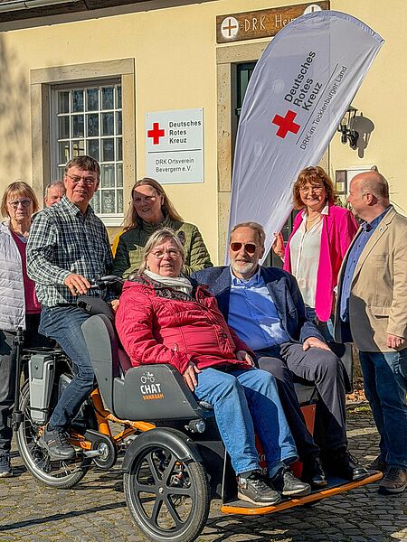 Rikscha-Projekt Ibbenbüren Gruppe von Personen vor dem Vereinsheim des DRK Ortsverein Bevergern e.V., darunter zwei ältere Menschen, die in einer Rikscha des Projekts ‚Radeln ohne Alter‘ sitzen. Im Hintergrund sind das DRK-Schild und eine Flagge des DRK Tecklenburger Land zu sehen.