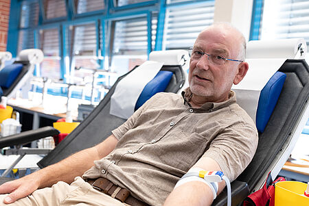 Ein älterer Mann mit Glatze und Brille liegt in einer Blutspendeliege und blickt zur Kamera. An seinem linken Arm ist ein Schlauch befestigt. Er trägt ein braunes, kurzärmeliges Hemd und eine helle Hose. Im Hintergrund sind weitere Blutspendeliegen und große Fenster zu sehen.
