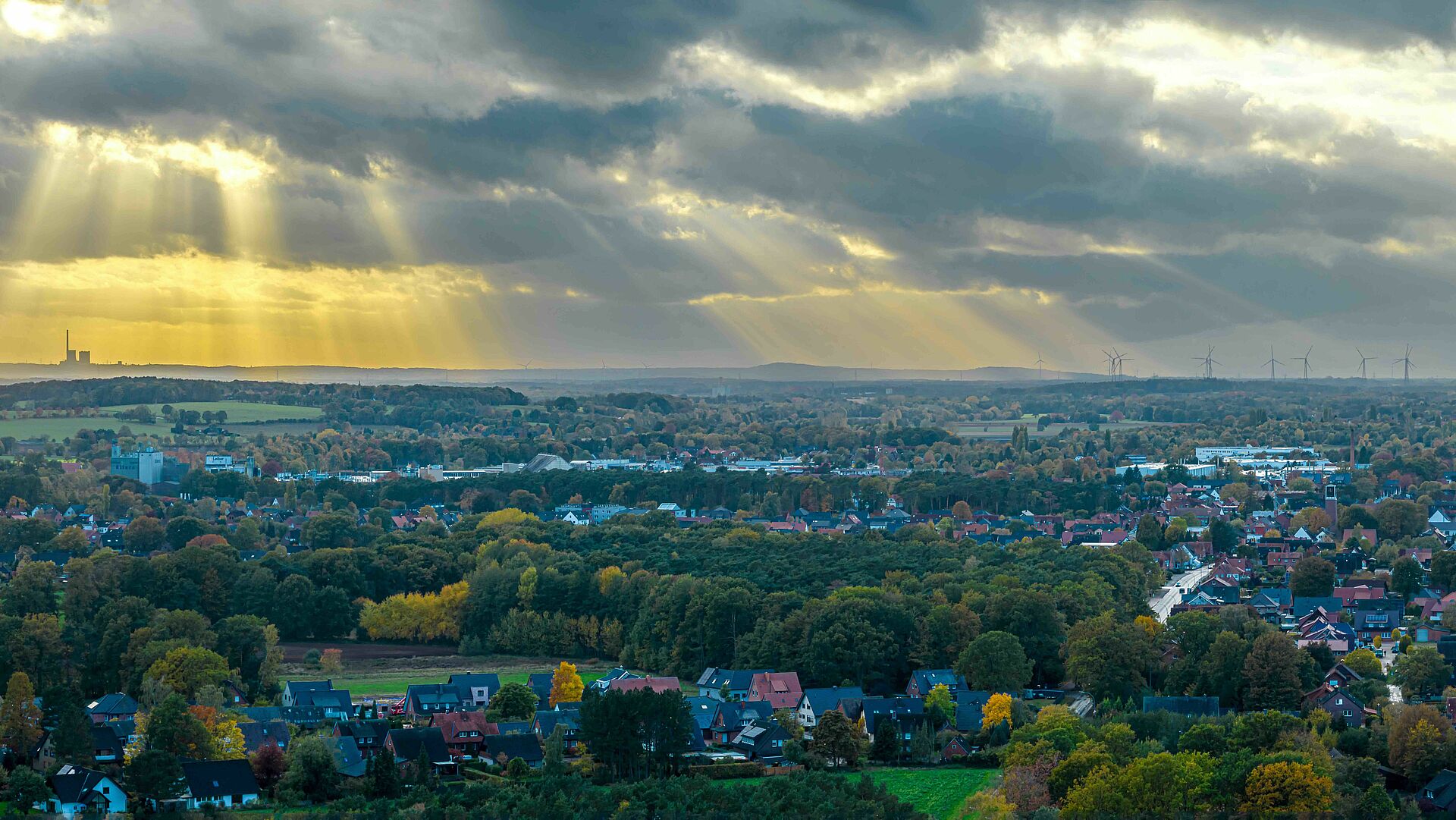 Header Startseite Ein Luftbild zeigt eine weite Landschaft des Tecklenburger Landes mit Häusern, Bäumen und Feldern, über denen Sonnenstrahlen durch die Wolken brechen. Am Horizont sind Industriebauten und Windräder zu erkennen.