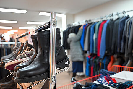 Kleidershop Lengerich In einem Secondhand- oder Sozialkaufhaus stehen vorne schwarze Lederstiefel und weitere Schuhe ordentlich auf einem Metallregal. Im Hintergrund hängt eine Vielzahl von Jacken und Kleidung auf einem Kleiderständer, während eine Person in heller Jacke danach greift. Die Szene wirkt wie ein sortierter Verkaufs- oder Ausgabebereich.