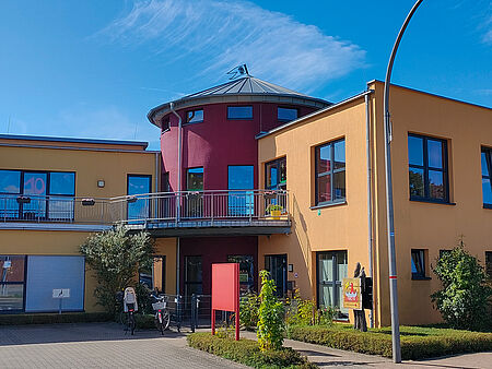 20250904_Besuchstag Modernes, zweigeschossiges Gebäude mit gelber und roter Fassade, großen Fenstern und einem runden Turm mit silbernem Dach. Vor dem Eingang stehen Fahrräder, Pflanzen und ein rotes Schild. Rechts am Gebäude ist ein buntes Logo mit einer Figur und dem Schriftzug „Kita Biberburg“ zu sehen. Der Himmel ist blau mit wenigen Wolken.