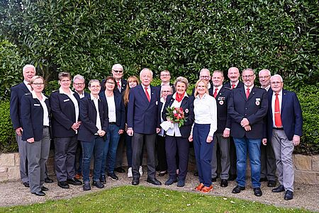 Bild 3 Verabschiedung Bernadette Gruppe von fünfzehn Personen in Uniformen des Deutschen Roten Kreuzes steht lächelnd in zwei Reihen vor einem grünen Buschhintergrund. Die Personen tragen dunkle Jacken, weiße Hemden und teilweise rote Krawatten oder Schals. Eine Person in der Mitte hält einen Blumenstrauß mit roten und rosafarbenen Rosen.