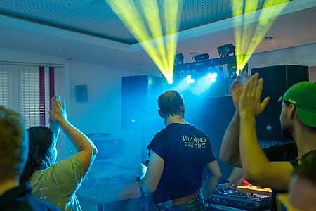 AuW_Disco Das Bild zeigt eine Diskoveranstaltung mit bunten Lichteffekten, bei der ein DJ Musik auflegt und Feiernde tanzen.