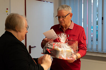 Verabschiedung Kammertöns Das Foto zeigt eine freundliche, persönliche Übergabesituation in einem Innenraum (vermutlich Büro oder Gemeinschaftsraum). Ein älterer Mann mit grauen Haaren und Brille trägt ein rotes Oberteil und hält einen liebevoll verpackten Geschenkkorb in den Händen. Der Geschenkkorb ist in transparenter Folie eingeschlagen und mit roten Schleifen dekoriert. Zusätzlich liest der Mann eine Karte, die offenbar zum Geschenk gehört. Ihm gegenüber steht eine ältere Frau mit Brille und kurzen grauen Haaren, die dunkel gekleidet ist. Sie lächelt und scheint das Geschenk gerade zu überreichen oder zu kommentieren. Die Körpersprache beider Personen wirkt wertschätzend und vertraut.