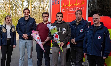 Azubis RW Eine junge Frau mit langen, hellen Haaren, die ein dunkles Jackett trägt. Daneben stehen fünf Männer, von denen zwei in Uniformen des DRK gekleidet sind. Die beiden Personen in der Mitte halten jeweils einen bunt gestalteten Konus, der typischerweise bei Geburtstagen oder Feiern im DRK-Bereich verwendet wird. Die Konusse tragen die Namen „DANI“ und „ELIAS“ sowie das Logo des DRK. Die Gruppe wirkt freundlich und posiert gemeinsam für das Foto. Im Hintergrund sind Bäume und der Rettungswagen zu sehen.
