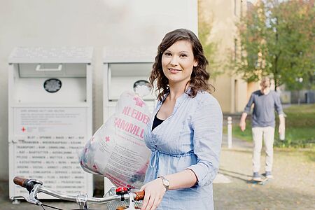 Symbolbild_Kleidercontainer Frau mit Fahrrad trägt einen Sack für die DRK-Kleidersammlung und steht vor zwei Sammelcontainern des Deutschen Roten Kreuzes im Außenbereich.