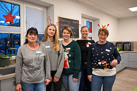 Adventszauberfest 2025 Fünf Frauen stehen lächelnd in einer weihnachtlich dekorierten Küche. Sie tragen Rentiergeweihe oder festliche Haarreifen sowie Namensschilder und weihnachtliche Pullover. Hinter ihnen hängen ein schwarzes Menübrett und rote Stern-Dekorationen an den Fenstern.