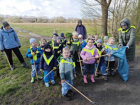 Besenparty Wiesenzwerge Eine Gruppe von Kindern in Warnwesten mit Greifzangen und Müllsäcken steht zusammen mit zwei erwachsenen Begleitpersonen auf einem Feldweg und sammelt Müll in der Natur. Alle wirken motiviert und zeigen ihre gesammelten Abfälle.