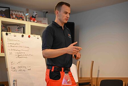 Praxisübung Ein Mann in dunkelblauem Poloshirt mit rotem Einsatzhosenteil steht vor einem Whiteboard und erklärt Inhalte. Auf dem Whiteboard sind handschriftliche Notizen zu Medikamenten zu sehen.