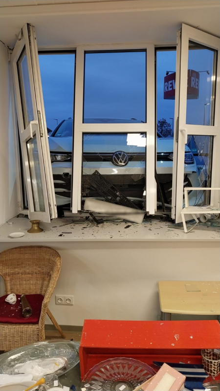 Unfall Lengerich Ein weißes VW-Auto ist durch ein Fenster in ein Gebäude gefahren. Die Scheiben sind zersplittert, Fahrzeugteile und Glasscherben liegen auf der Fensterbank und im Raum. Möbel wie ein Korbstuhl und ein kleiner Tisch stehen zwischen den Trümmern.