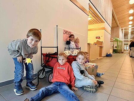 Aktion_Spendensammlung_Schatzkiste Eine Gruppe von Kindern sitzt und steht in einem hellen Flur. Vor ihnen steht ein roter Bollerwagen, daneben ein großer Karton mit gespendeten Lebensmitteln. Hinter dem Wagen hängt ein handgemaltes Schild mit der Aufschrift „Spende für die Tafel“. Die Kinder schauen in Richtung Kamera.