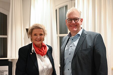 JHV 2025 Zwei Personen stehen nebeneinander in einem hellen Innenraum. Links steht eine Frau mit hellem, kurzen Haar, dunkelblazer, weißer Bluse und rotem Halstuch. Rechts steht ein Mann mit kurzem, hellem Haar, Brille und grauem Anzug über einem hellblauen Hemd. Beide lächeln in die Kamera. Im Hintergrund hängen helle Vorhänge und es sind Fenster zu sehen.