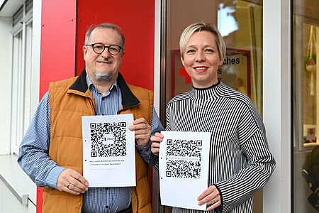Social Media - Chancen und Risiken für Menschen mit Beeinträchtigung Zwei Personen stehen vor einem Gebäude mit rotem Element und einem DRK-Logo im Hintergrund. Sie halten jeweils ein Blatt Papier mit großen QR-Codes in die Kamera. Die linke Person trägt eine Brille, ein kariertes Hemd und eine braune Weste, die rechte Person trägt einen weiß-dunkelblau gestreiften Pullover. Beide lächeln und präsentieren die Ausdrucke frontal.