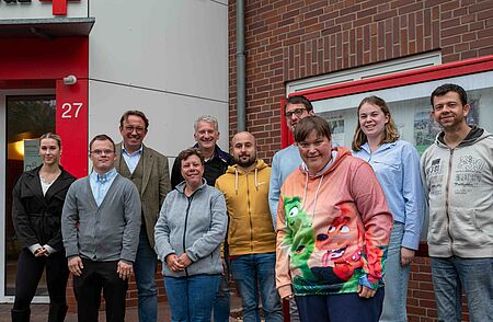 Klient:innenbeirat trifft den Bürgermeister Mitglieder des Klientenbeirats posieren gemeinsam vor einem Backsteingebäude mit der Hausnummer 27. Die Gruppe ist in zwei Reihen aufgestellt und lächelt in die Kamera; die Stimmung ist freundlich und locker.