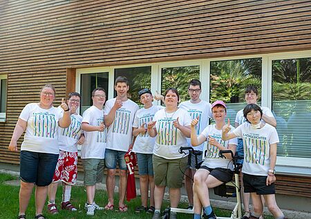 Mieter_innen Lachgesichter Gruppe von neun Personen, die vor einem Holzgebäude mit großen Fenstern stehen. Alle tragen weiße T-Shirts mit der Aufschrift '10 Jahre Lachgesichter' und bunte Muster. Einige zeigen den Daumen nach oben, andere lächeln in die Kamera. Eine Person sitzt in einem Rollstuhl, eine weitere trägt eine rote Basecap. Im Hintergrund sind grüne Sträucher und Bäume zu sehen.